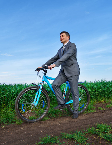 zakenman met een fiets poseert in een tarweveld, groen gras en blauwe lucht als achtergrond - Foto, afbeelding