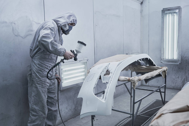 Car painting and automobile repair service. Auto mechanic in white overalls paints car with airbrush pulverizer in paint chambe - Fotografie, Obrázek