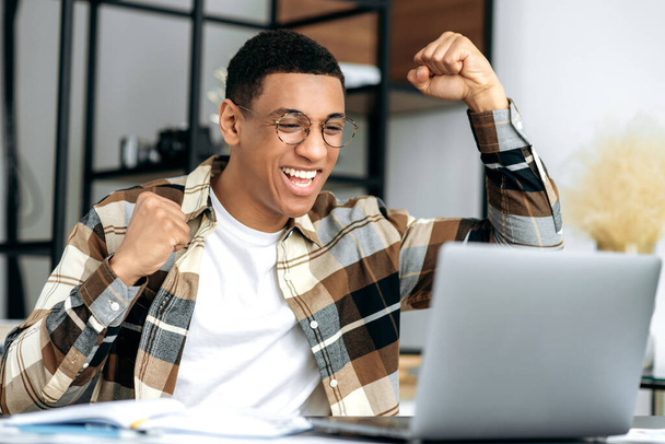 Happy positive successful hispanic young man, stylishly dressed, office worker, freelancer or designer sits at work desk, gesturing with his hands rejoicing in victory, success, good deal, big profit - Photo, Image