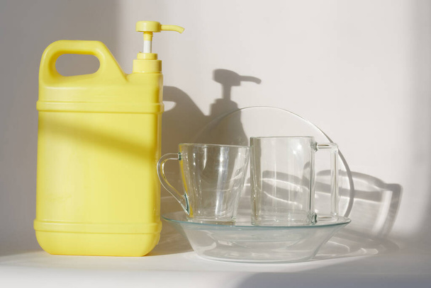 Keltainen muovisäiliö pumpulla annostelija ja puhdistus desinfiointiaine neste seisoo vieressä puhdas läpinäkyvä ruokalaji, auringonsäteet. Keittiön desinfiointi ja puhdistus käsite. lähikuva - Valokuva, kuva