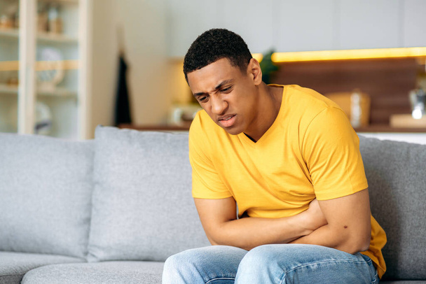 Hispanic frustrated unhealthy guy, sitting at home on the sofa, holding his stomach with his hands, suffering from stomach pain, stomach cramps, problems with the digestive tract, needs treatment - Foto, Imagen