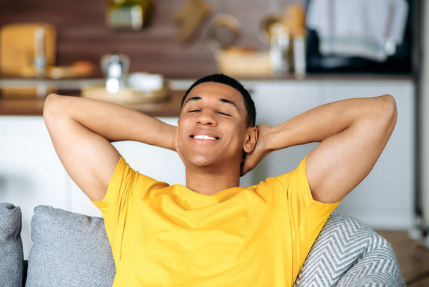 Tevreden jonge latino man ontspant zich op een bank in de woonkamer, glimlachend. Een knappe latino die thuis aan het chillen is, luie vrije tijd heeft, droomt over vakanties, zijn handen achter het hoofd gooit - Foto, afbeelding