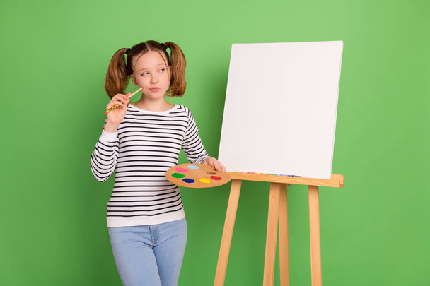Photo of thoughtful pretty small schoolgirl dressed striped sweater drawing looking canvas empty space isolated green color background - Fotó, kép