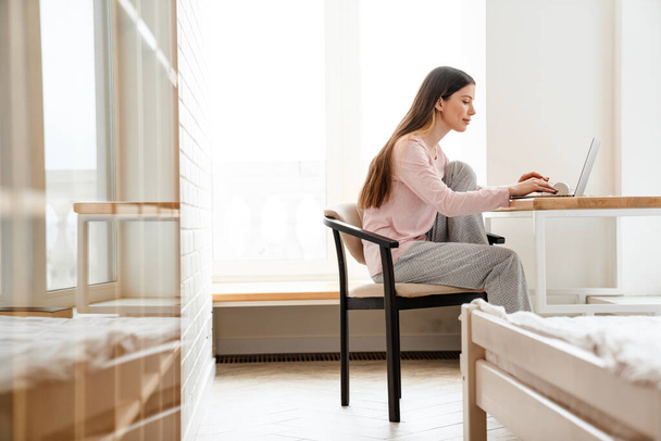 Happy young white woman wearing lounge clothes sitting at the table with laptop computer, studying, video calling - 写真・画像