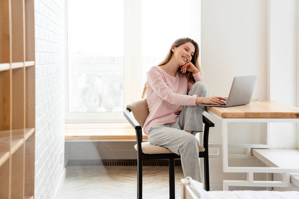 Glückliche junge weiße Frau in Lounge-Kleidung sitzt am Tisch mit Laptop-Computer, studiert, Videotelefonie - Foto, Bild