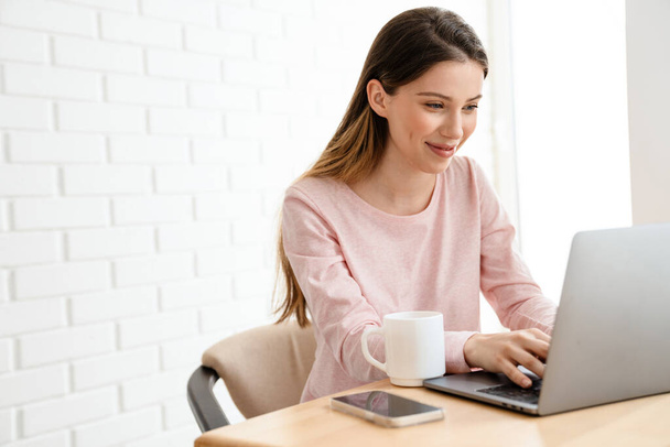 Happy young white woman wearing lounge clothes sitting at the table with laptop computer, studying, video calling - Photo, Image