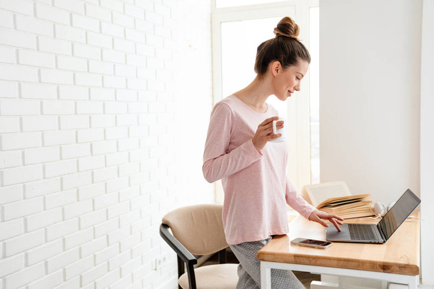 Happy young white woman wearing lounge clothes standing at the table with laptop computer, studying, video calling, holding mug - Zdjęcie, obraz
