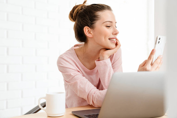 Happy young white woman wearing lounge clothes sitting at the table with laptop computer, studying, video calling, holding mobile phone - 写真・画像