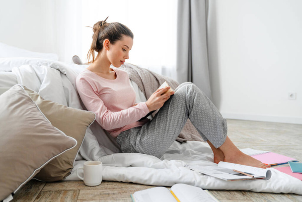 Young smiling white woman reading book studying sitting on a floor of her bedroom on a blanket - Photo, Image