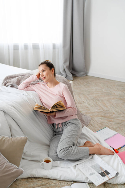 Young white woman reading book studying sitting on a floor of her bedroom on a blanket - Foto, immagini