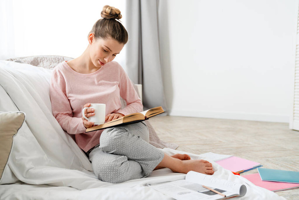 Smiling young white woman in lounge wear sitting on a floor in the bedroom, reading book, holding cup - Фото, зображення
