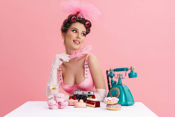 Young woman wearing curlers holding feather while sitting at table isolated over pink background - Foto, imagen