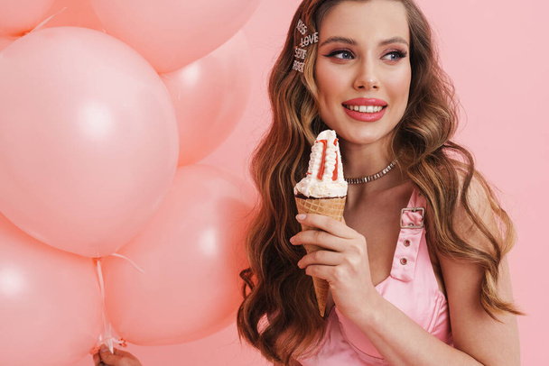 Young smiling woman eating ice cream while posing with balloons isolated over pink background - Фото, зображення