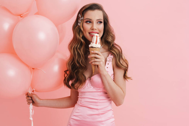 Young smiling woman eating ice cream while posing with balloons isolated over pink background - Zdjęcie, obraz
