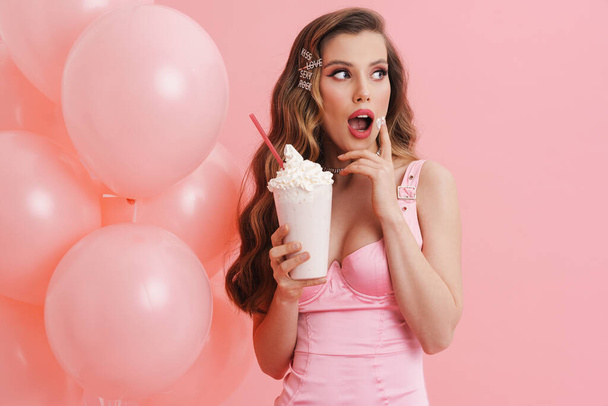 Young surprised woman eating cream while posing with balloons and milkshake isolated over pink background - Foto, Imagen