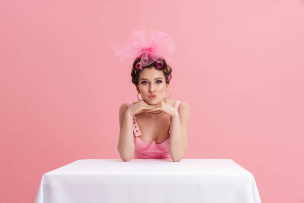 Young woman wearing curlers making kiss lips while sitting at table isolated over pink background - Foto, imagen