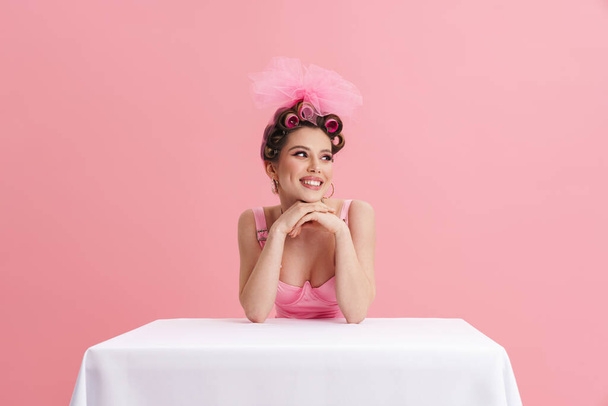 Young woman wearing curlers smiling at camera while sitting at table isolated over pink background - Fotoğraf, Görsel