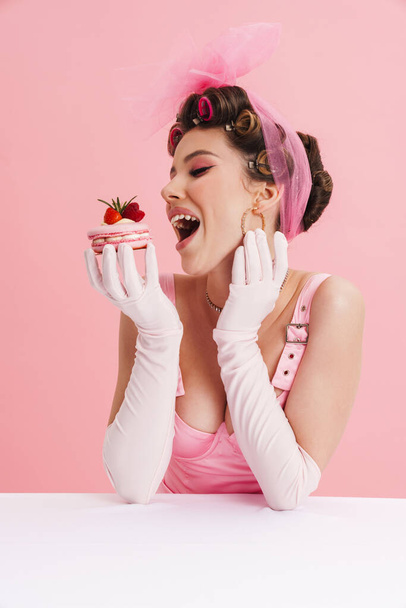 Smiling woman wearing gloves eating cake while sitting at table isolated over pink background - Fotó, kép