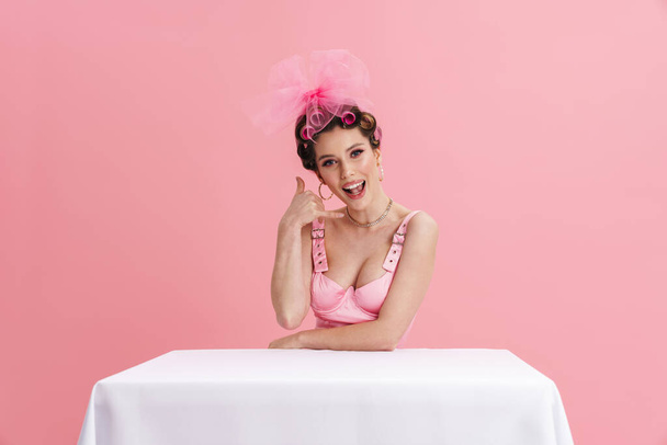 Young woman wearing curlers showing handset gesture while sitting at table isolated over pink background - Foto, Imagen