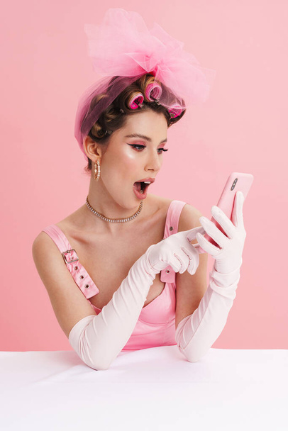 Young smiling white woman barbie in pink dress sitting at the table isolated over background, holding mobile phone - Foto, Bild