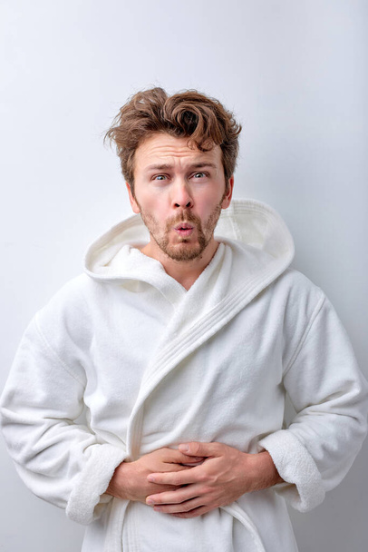 Young bearded man wearing bathrobe with hand on stomach because indigestion - 写真・画像