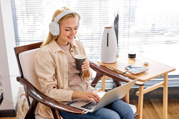 Blonde Caucasian Female Sits Working On Laptop, In Headset, Using Humidifier - Photo, Image
