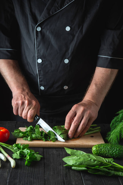 Een chef-kok bereidt een spinaziesalade in een keuken van het restaurant. Sluiten van de handen van een kok die groenten snijdt met een mes op een snijplank. - Foto, afbeelding