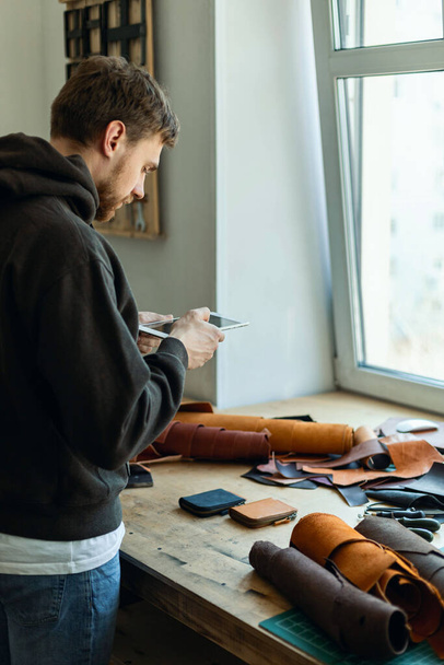 Top view male tanner use tablet at leather workshop. Craftsman creating online advertising promotion - Fotografie, Obrázek