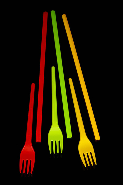Пластиковые одноразовые столовые приборы: желтый, зеленый и красный (цвета флага Литвы) вилки и соломинки для коктейлей изолированы на черном фоне - Фото, изображение