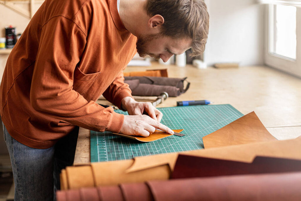 Male tanner making cutting scheme of bag working at leather workshop. Handcrafted creating - Fotoğraf, Görsel