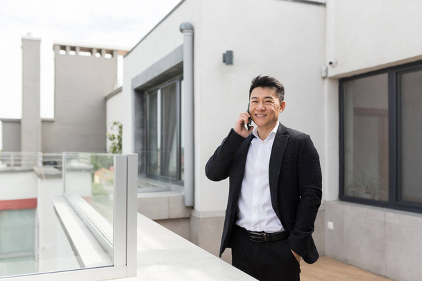 Freelancer Asian businessman in business suit enjoys the phone standing on the balcony terrace of a modern office center successful and happy - Φωτογραφία, εικόνα