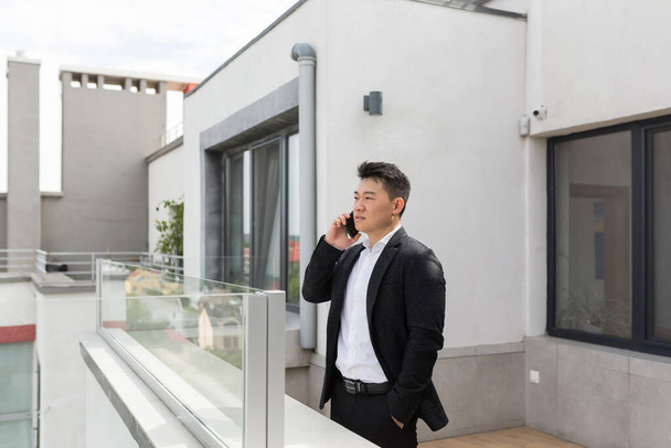 Ελεύθερος επαγγελματίας Ασιάτης επιχειρηματίας με κοστούμι επιχειρήσεων απολαμβάνει τηλέφωνο στέκεται στη βεράντα μπαλκόνι του σύγχρονου κέντρου γραφείων - Φωτογραφία, εικόνα