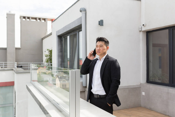 Freelancer Asian businessman in business suit enjoys phone standing on balcony terrace of modern office center - Fotografie, Obrázek