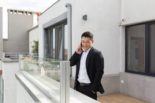 Freelancer Asian businessman in business suit enjoys the phone standing on the balcony terrace of a modern office center successful and happy - Φωτογραφία, εικόνα