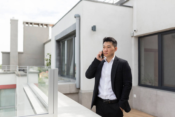 Freelancer Asian businessman in business suit enjoys phone standing on balcony terrace of modern office center - Φωτογραφία, εικόνα