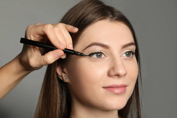 Artist applying black eyeliner onto woman's face on grey background, closeup - Zdjęcie, obraz