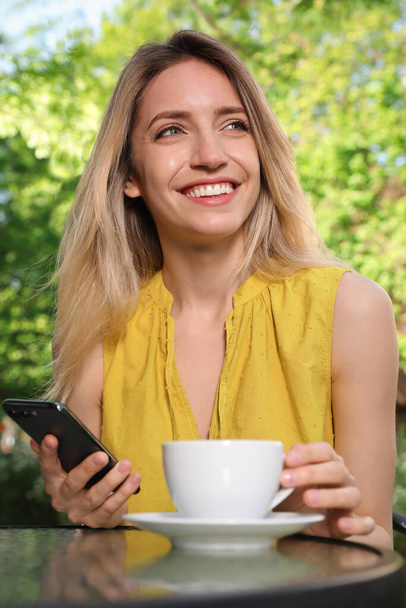 Happy young woman with cup of coffee and smartphone enjoying early morning in outdoor cafe - Foto, Imagem