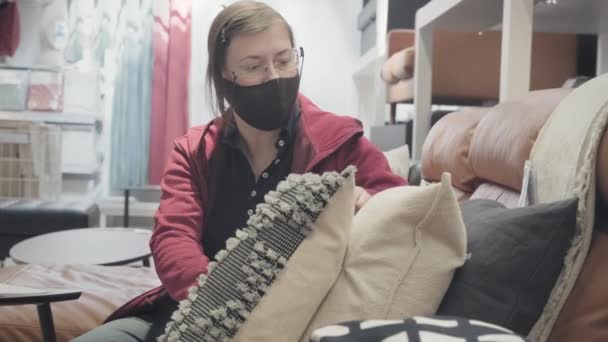 Mujer en contra de la máscara de virus se sienta en el sofá en la tienda y selecciona almohadas suaves - Metraje, vídeo