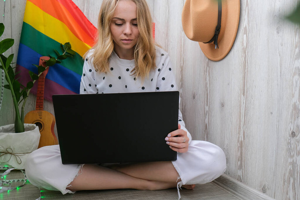 若い千年ヒッピー女性がラップトップを使ってバルコニーに座っています。背景にLGBTQの虹の旗。オンライン教育授業料。家から仕事だ。近代事務所. - 写真・画像