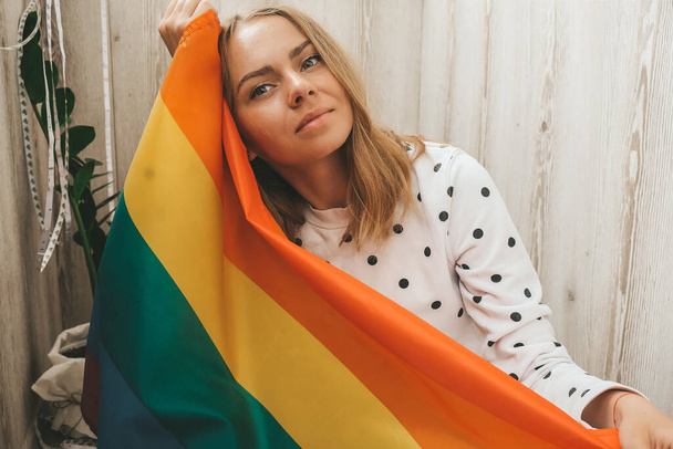 Young blonde millennial hippie woman with rainbow LGBTQ flag at home balcony. Peace and freedom the symbol of LGBT, spending and sharing loving time. Support LGBTQ community. - Photo, Image