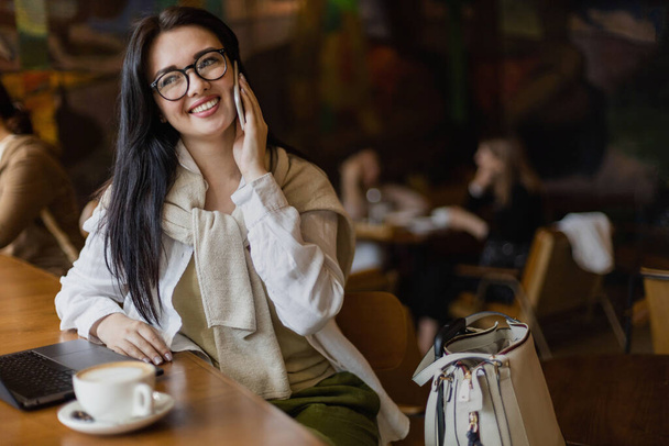 Pretty young business woman talking smartphone sitting at table in cafe with laptop and coffee cup - Photo, Image