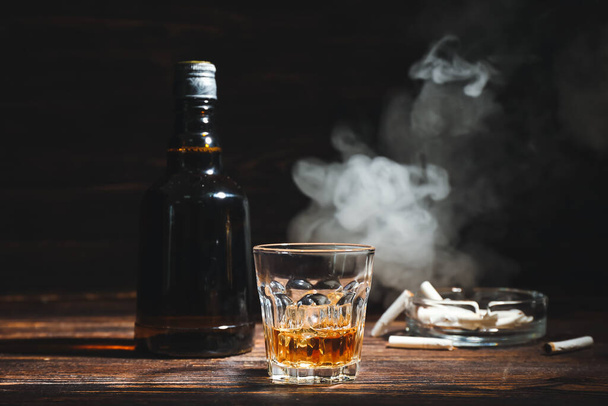 Бутылка и стакан холодного виски, сигареты на деревянном столе на тёмном фоне - Фото, изображение