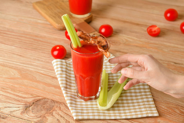 Female hand with glass of bloody mary cocktail garnished with bacon and celery on wooden background - Photo, Image