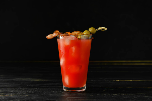Glass of bloody mary cocktail garnished with bacon and olives on dark wooden background - Фото, зображення