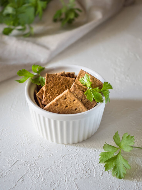 Natural flat rye mini-breads of a square shape in a white bowl on a light concrete background. Textile and parsley in the background. Delicious organic and quick snacks for diet meals - Foto, afbeelding