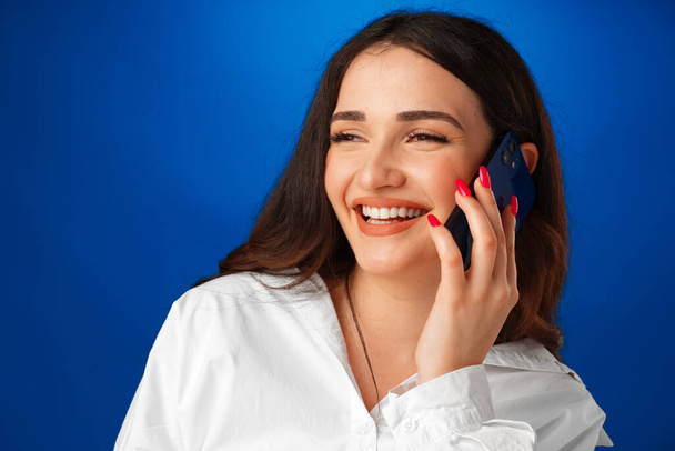 jong brunette vrouw praten op de telefoon tegen blauw achtergrond - Foto, afbeelding