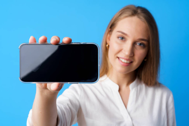 Beautiful young woman showing smartphone screen against blue background - Fotoğraf, Görsel