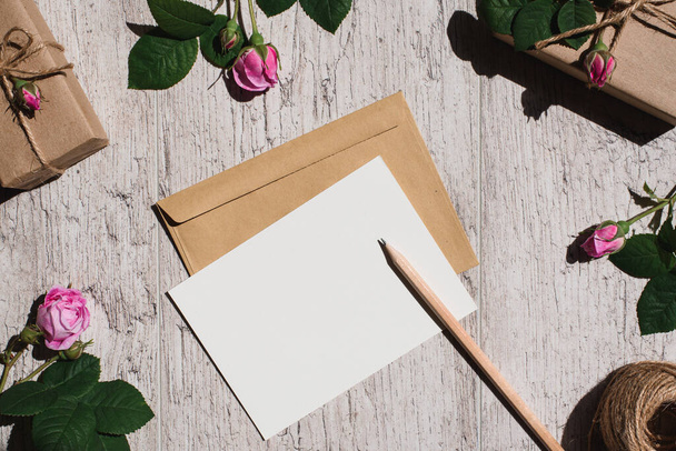 Mockup blanke weiße Grußkarte oder Einladung, kraftbrauner Umschlag, rosa Rosenblumen und Geschenkschachteln auf grauem Hintergrund. Flache Lage, Draufsicht. Kopierraum - Foto, Bild