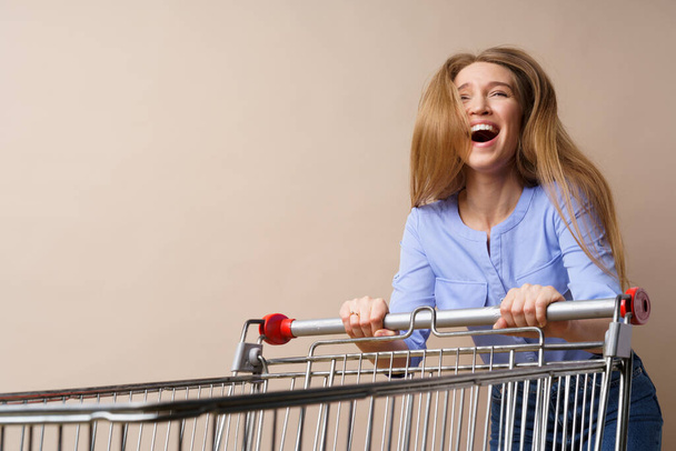 Young cheerful woman with empty shopping cart on beige background - Fotografie, Obrázek