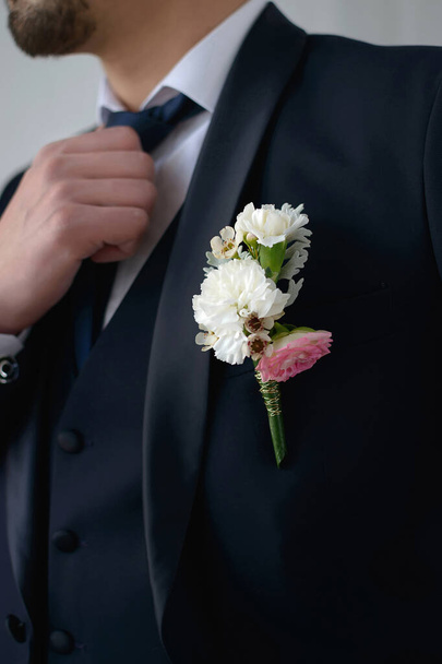 Ο γαμπρός με σκούρο μπλε κοστούμι με μπουτονιέρα προσαρμόζει τη γραβάτα του κοιτάζοντας στο πλάι σε κοντινό πλάνο. - Φωτογραφία, εικόνα